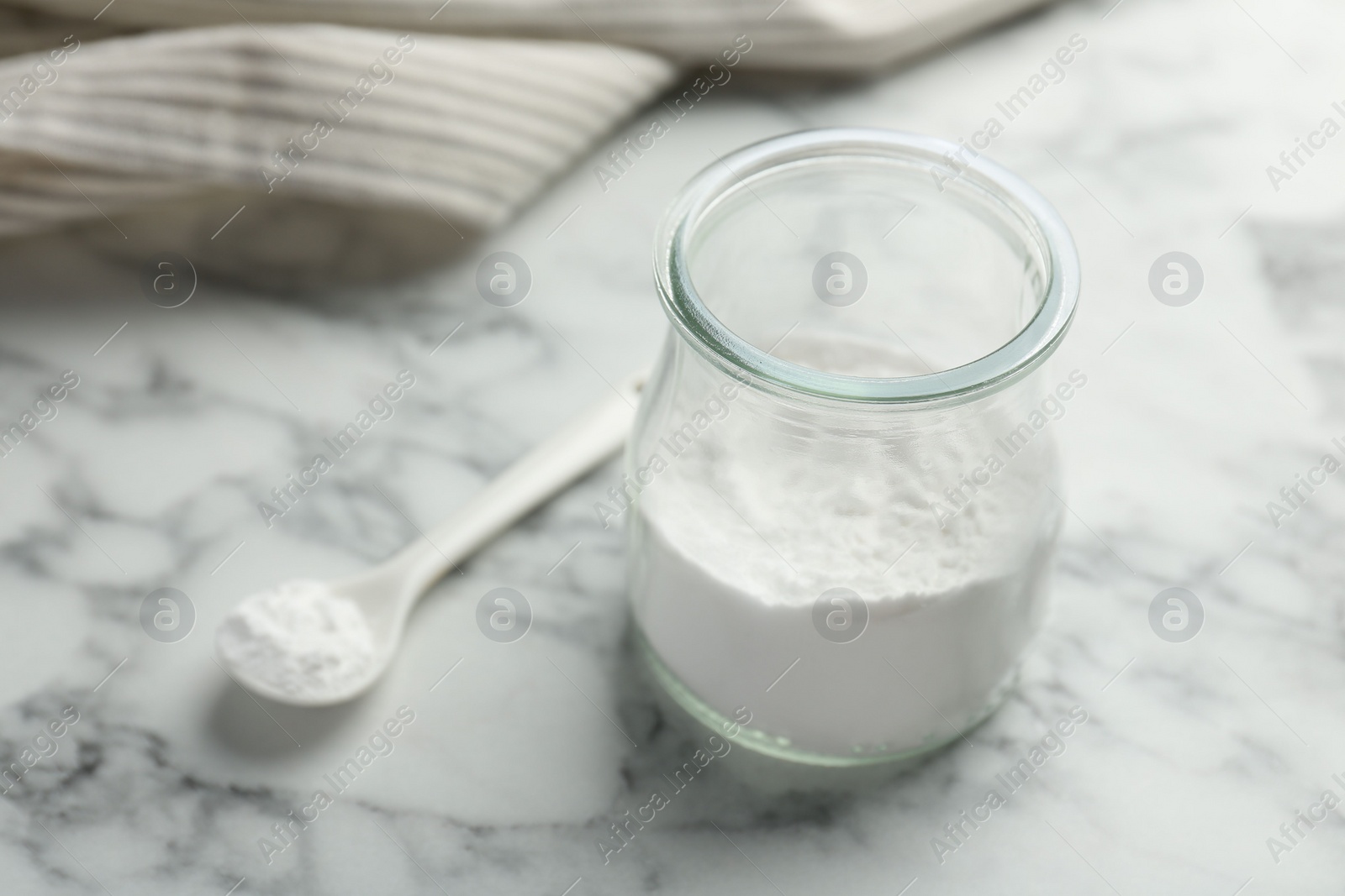 Photo of Baking powder in jar and spoon on white marble table, closeup. Space for text