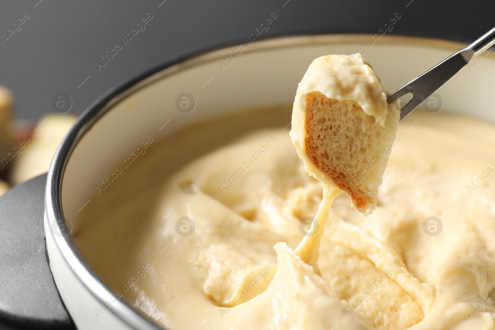 Photo of Dipping piece of bread into fondue pot with melted cheese on grey background, closeup