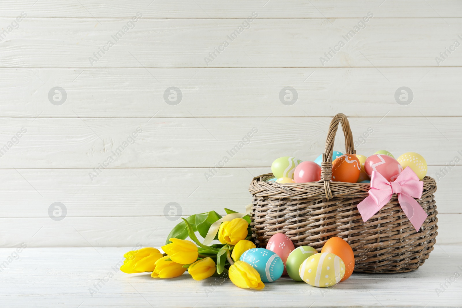 Photo of Wicker basket with painted Easter eggs and spring flowers on table, space for text