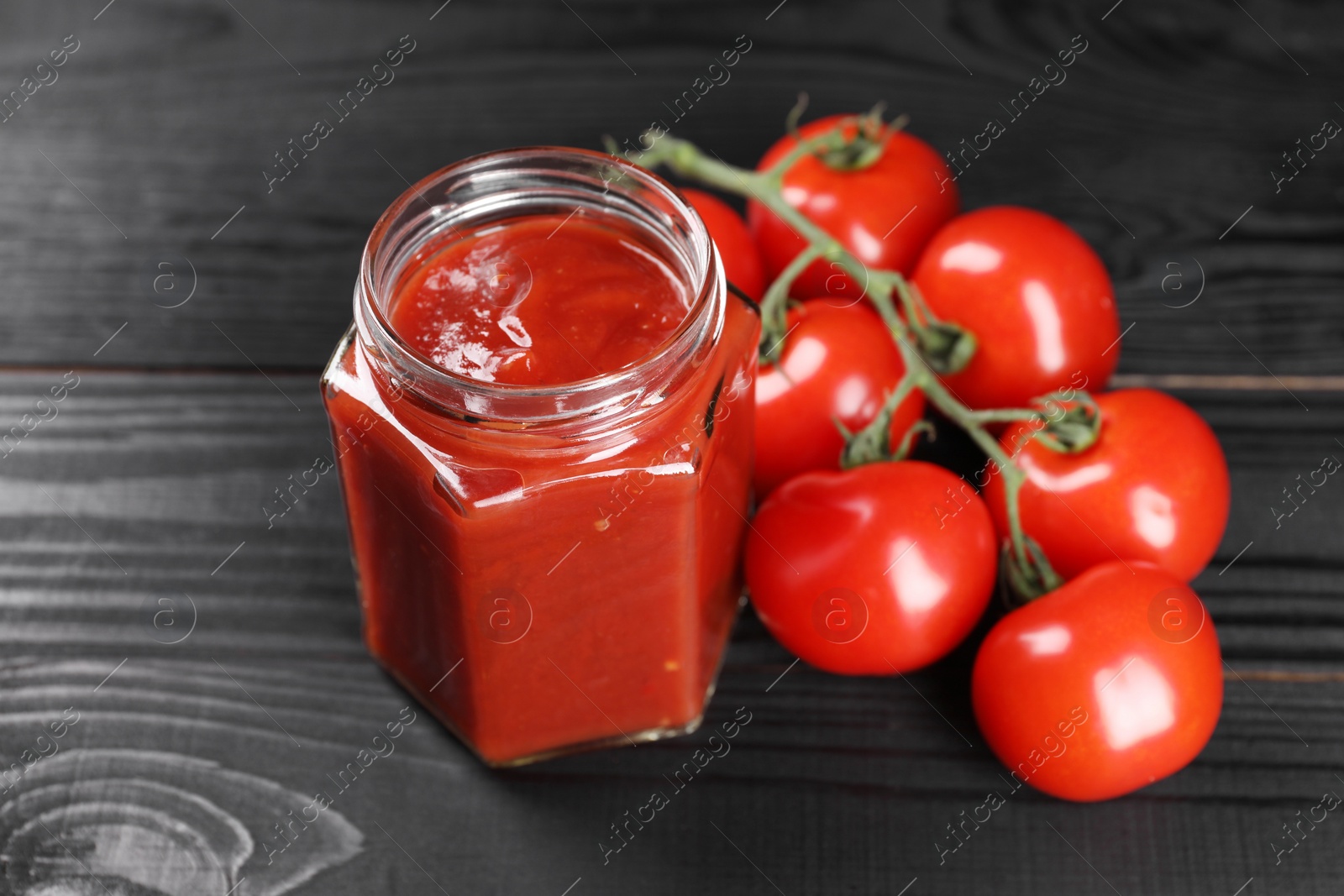 Photo of Delicious ketchup in jar and tomatoes on black wooden table, closeup. Tomato sauce