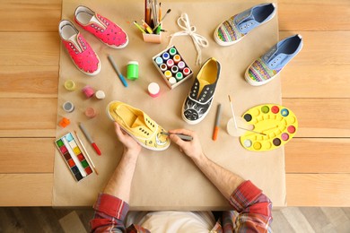 Photo of Man painting on sneaker at wooden table, top view. Customized shoes