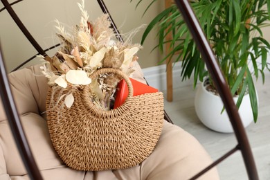 Stylish beach bag with beautiful bouquet of dried flowers and book on papasan chair indoors