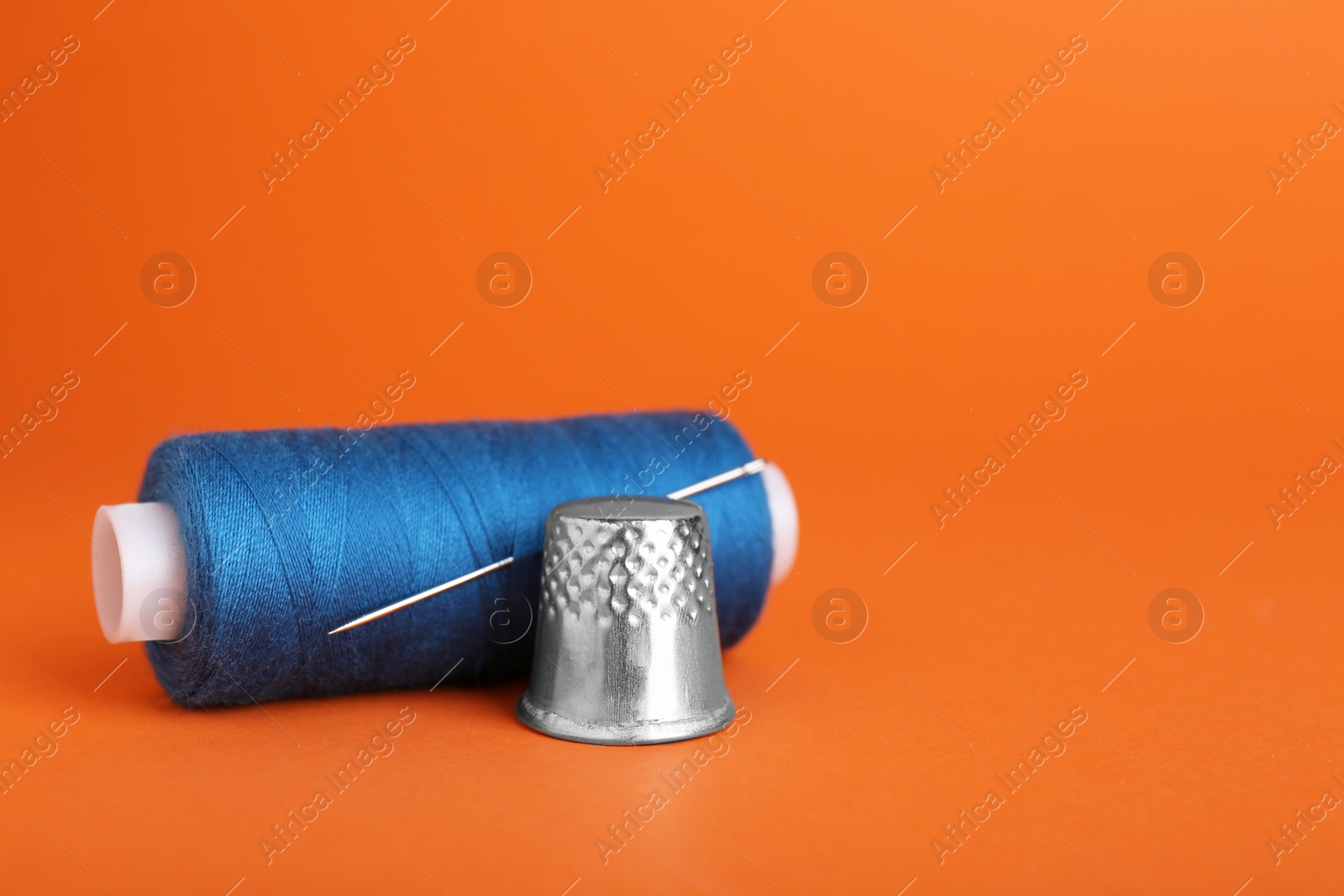 Photo of Thimble and spool of blue sewing thread with needle on orange background, closeup. Space for text