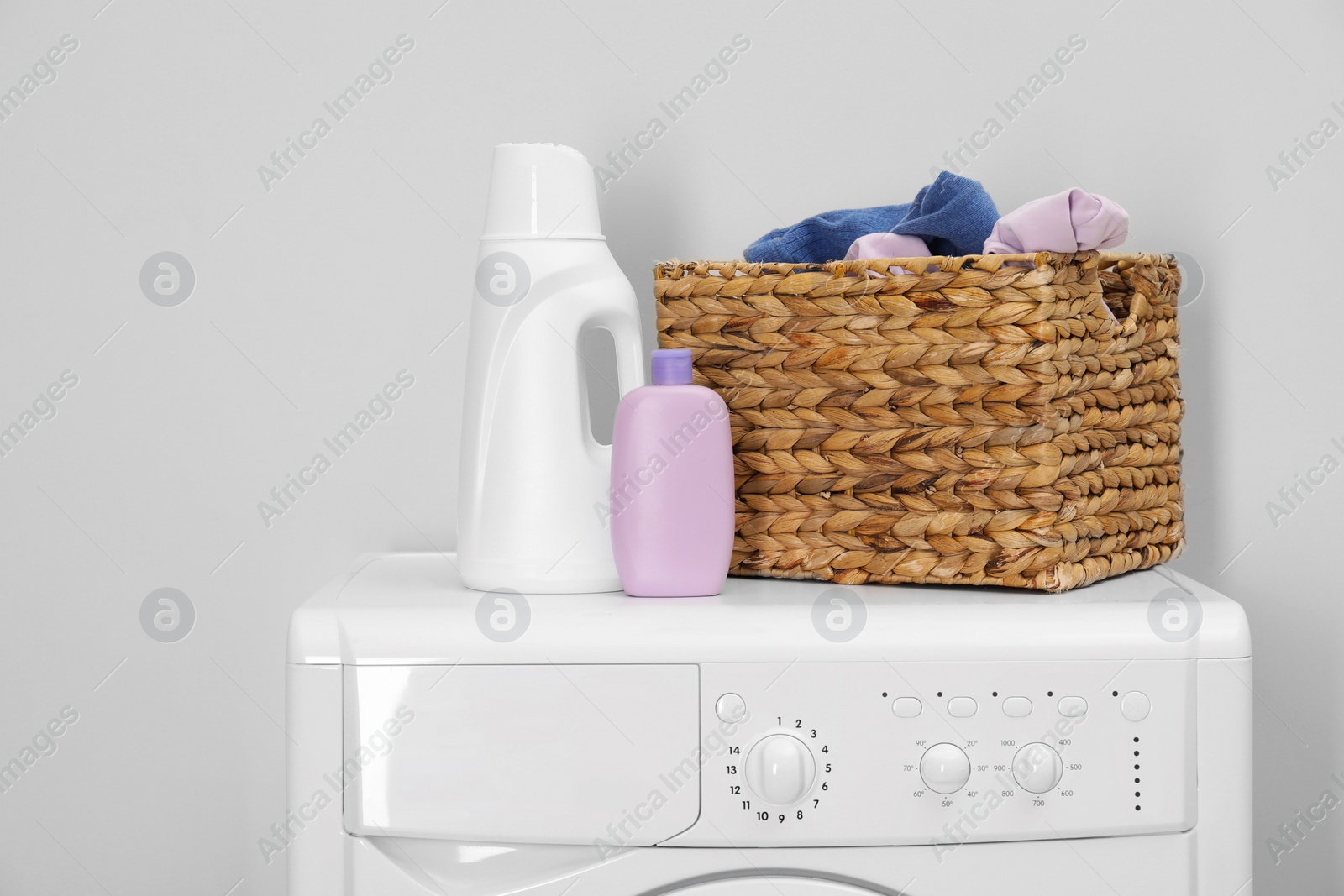 Photo of Baby clothes in wicker basket and laundry detergents on washing machine near light wall, space for text