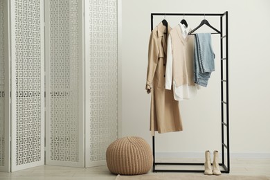 Photo of Rack with different stylish women's clothes, boots, pouf and folding screen indoors