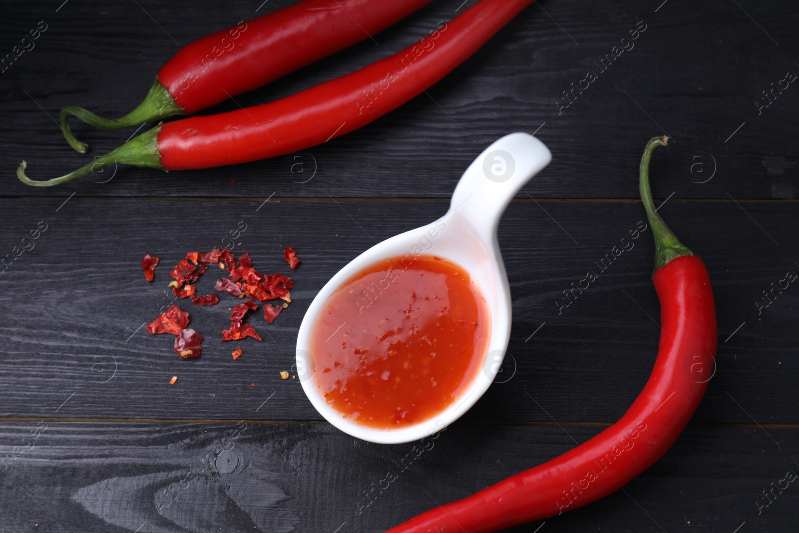 Photo of Spicy chili sauce in spoon and peppers on black wooden table, flat lay