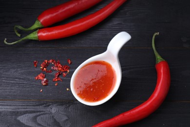 Photo of Spicy chili sauce in spoon and peppers on black wooden table, flat lay