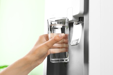 Photo of Woman filling glass from water cooler indoors, closeup. Refreshing drink