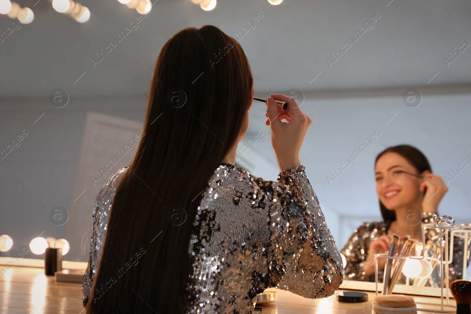 Photo of Young woman applying make up near illuminated mirror indoors