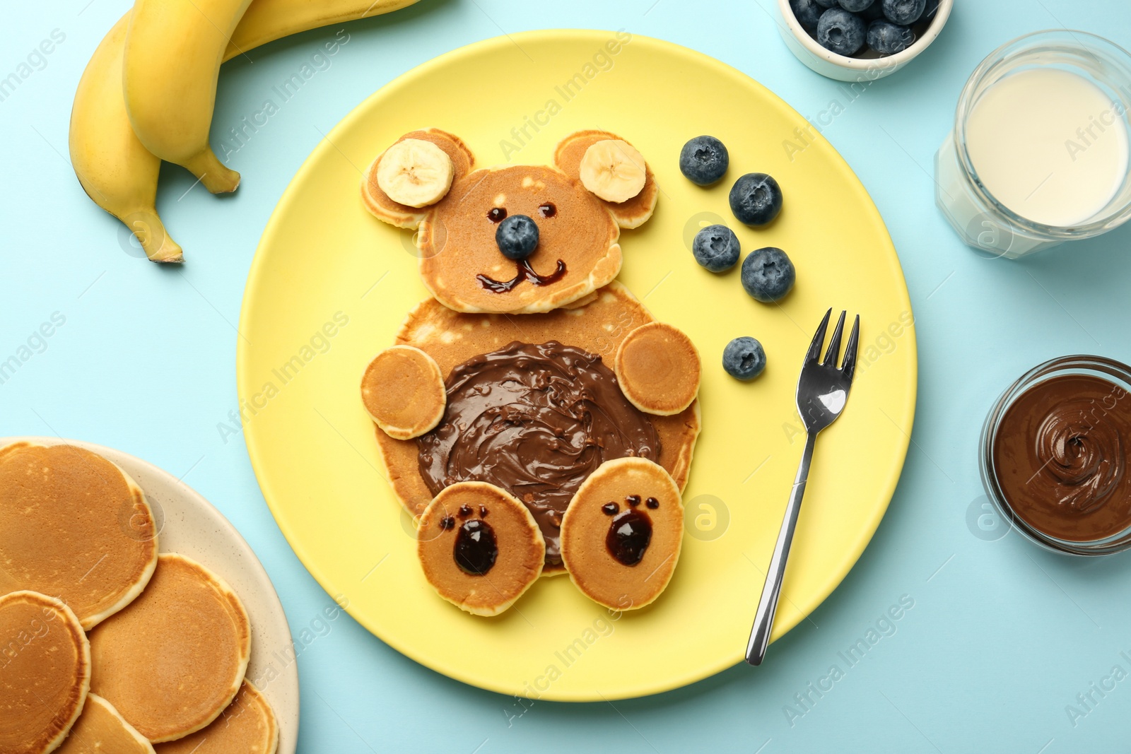 Photo of Creative serving for kids. Plate with cute bear made of pancakes, blueberries, bananas and chocolate paste on light blue table, flat lay