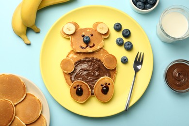Creative serving for kids. Plate with cute bear made of pancakes, blueberries, bananas and chocolate paste on light blue table, flat lay
