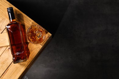 Whiskey with ice cubes in glass and bottle on wooden crate against black background, top view. Space for text