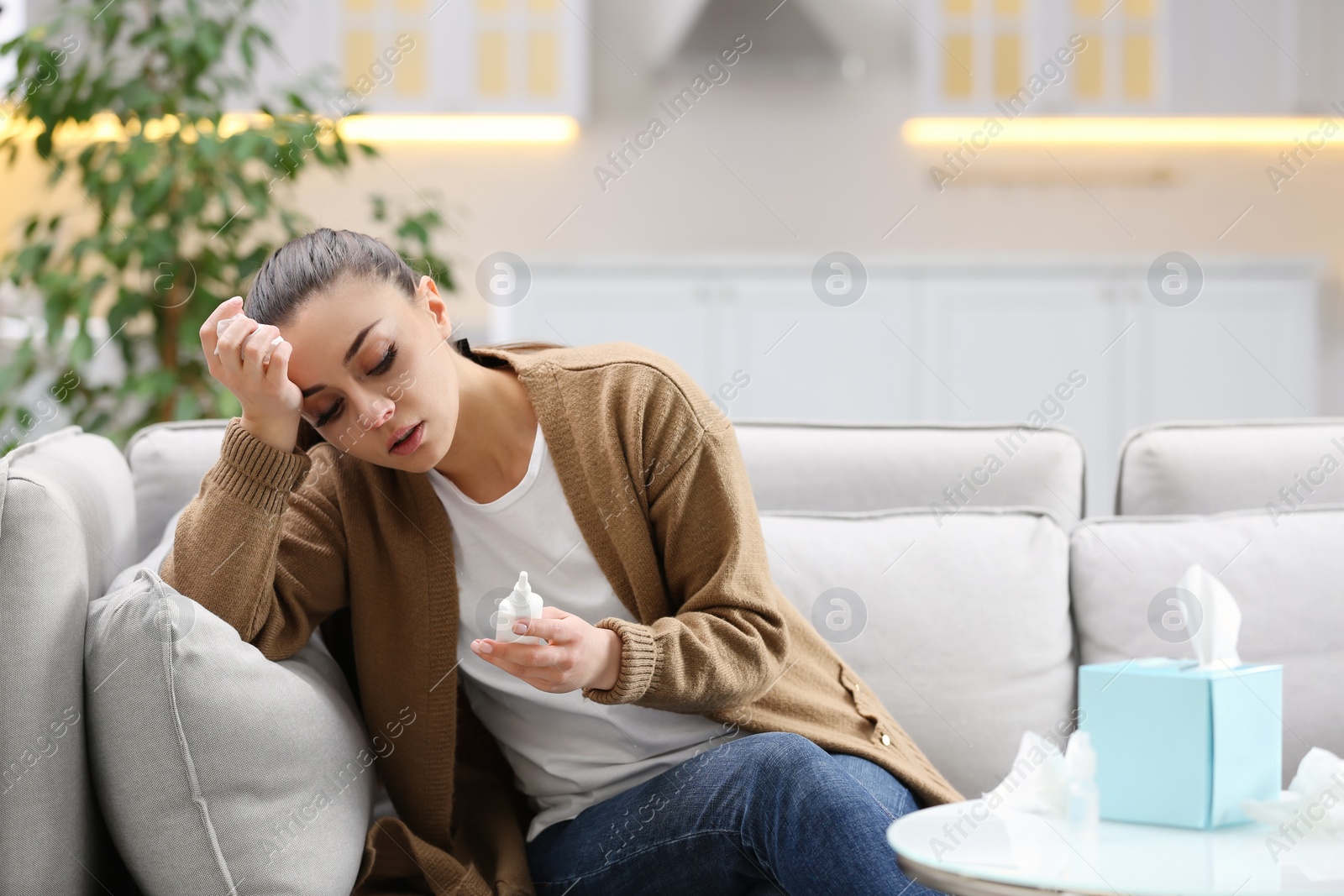 Photo of Ill woman with nasal spray and box of paper tissues on sofa at home