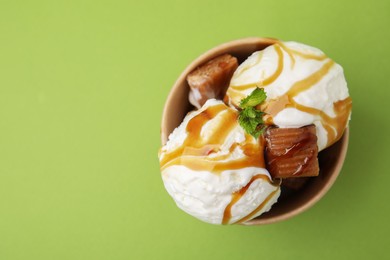 Photo of Scoops of tasty ice cream with mint, caramel sauce and candies in paper cup on green background, top view. Space for text