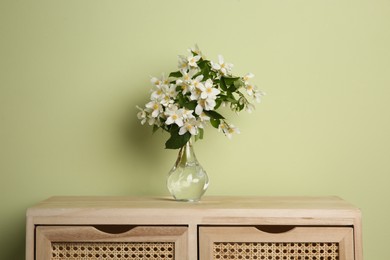 Bouquet of beautiful jasmine flowers in vase on wooden commode near light green wall