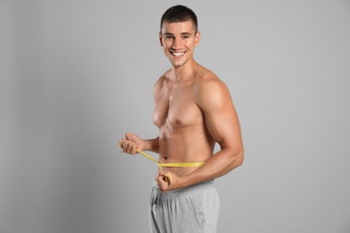 Young man with slim body using measuring tape on grey background