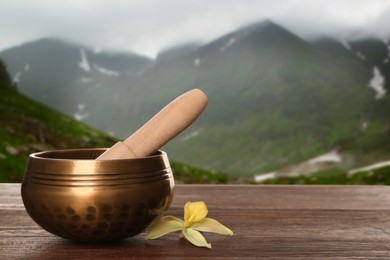 Image of Golden singing bowl, mallet and flower on wooden table against mountain landscape. Space for text 