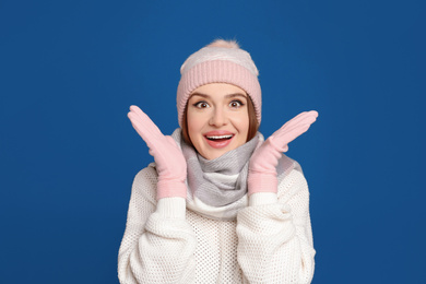 Photo of Excited woman wearing warm sweater, gloves, scarf and hat on blue background. Winter season