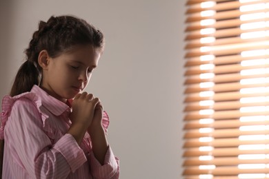 Photo of Cute little girl with hands clasped together praying near window. Space for text
