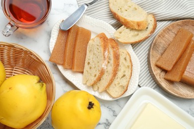 Delicious quince paste, bread, butter, cup of tea and fresh fruits on white marble table, flat lay