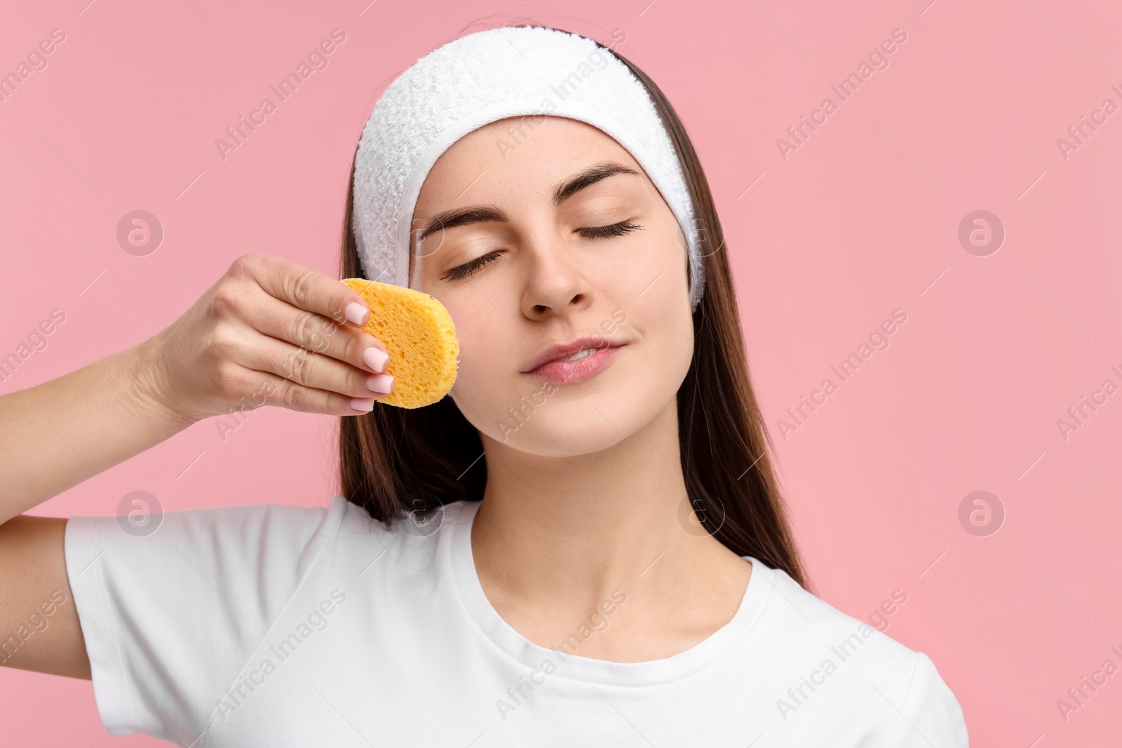 Photo of Young woman with headband washing her face using sponge on pink background