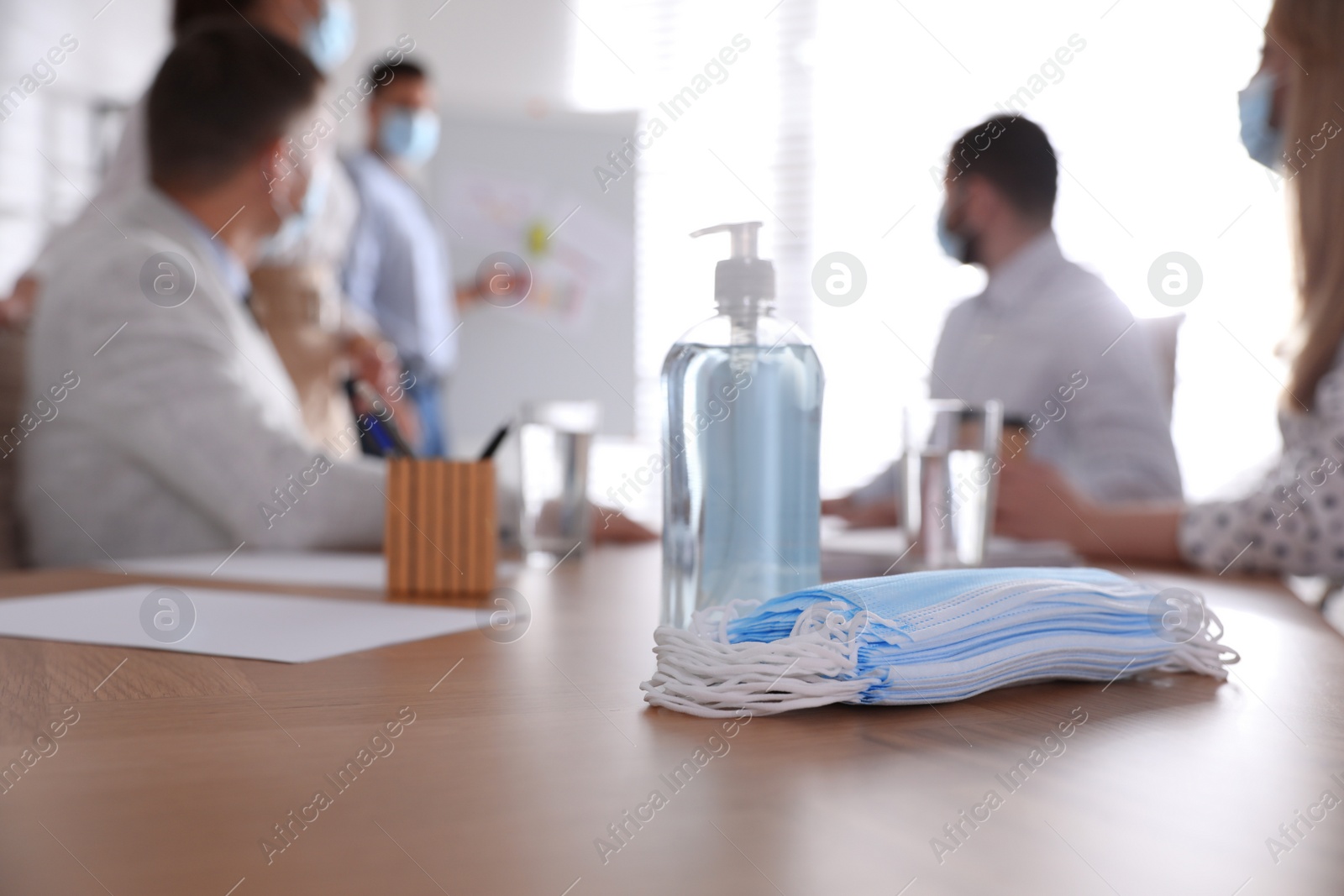 Photo of Protective masks, hand sanitizer and blurred view of coworkers on background. Business meeting during COVID-19 pandemic