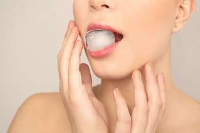 Young woman holding ice cube in mouth on light background, closeup