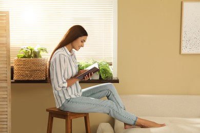 Photo of Beautiful young woman reading book on stool at home