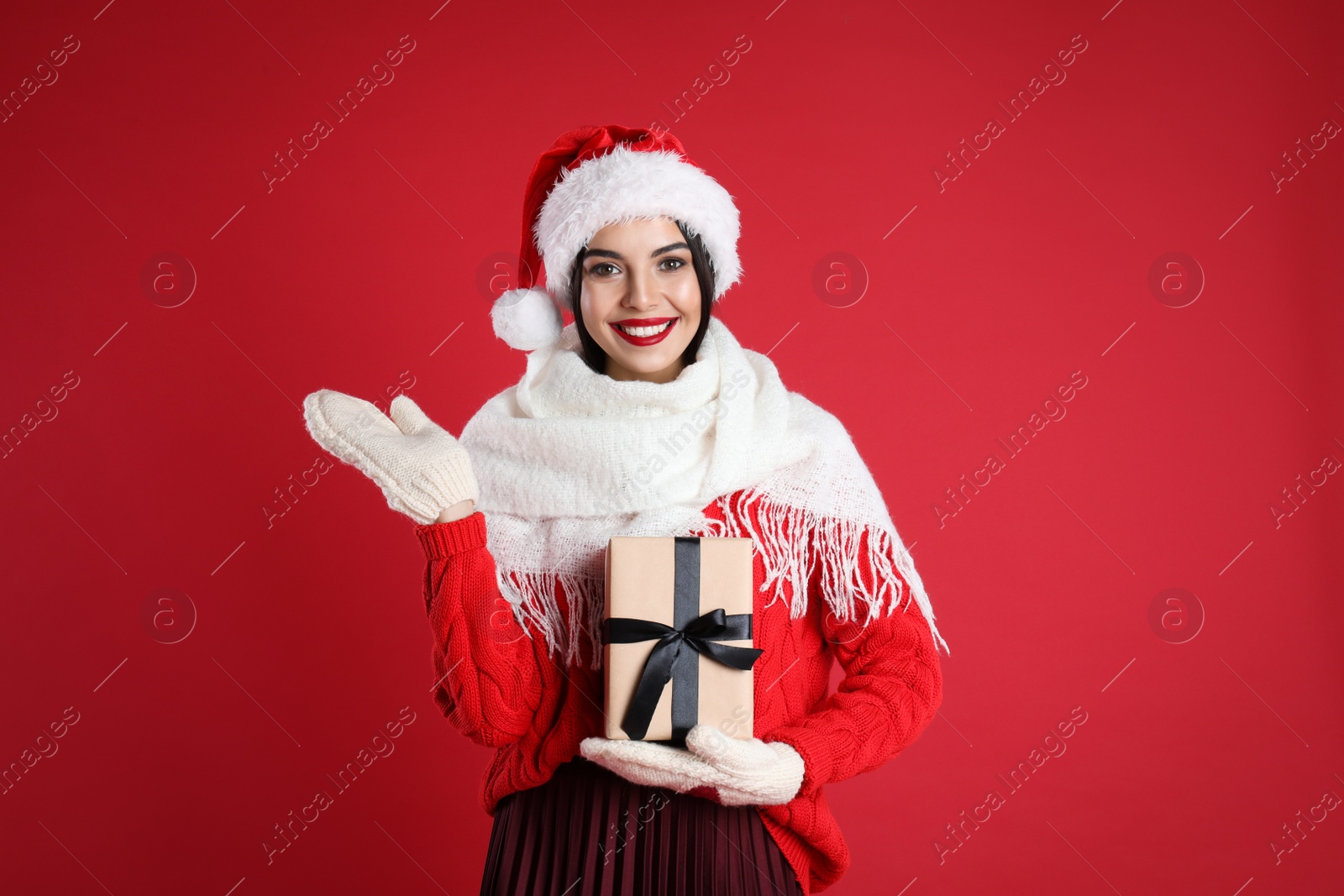 Photo of Woman in Santa hat, knitted mittens, scarf and sweater holding Christmas gift on red background