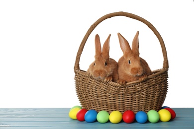 Photo of Cute fluffy bunnies in wicker basket near Easter eggs on light blue wooden table. Space for text
