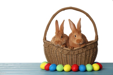 Cute fluffy bunnies in wicker basket near Easter eggs on light blue wooden table. Space for text