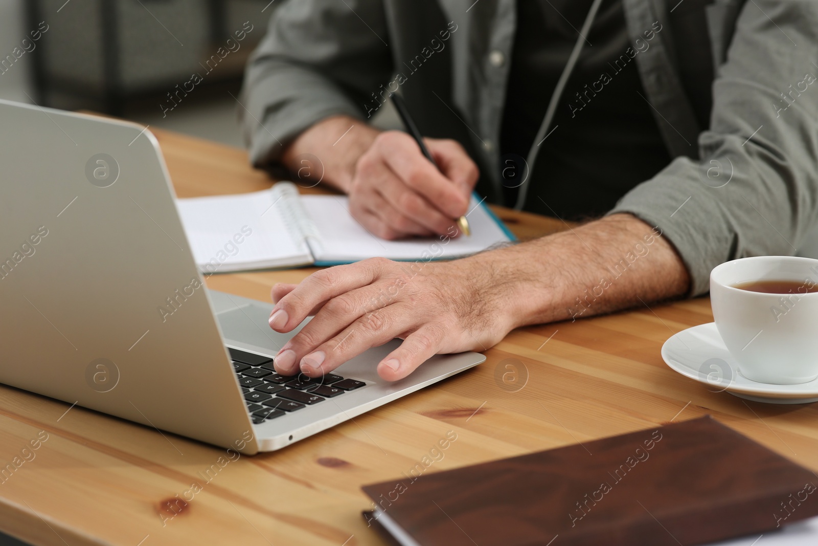 Photo of Man with laptop and notebook learning at wooden table indoors, closeup