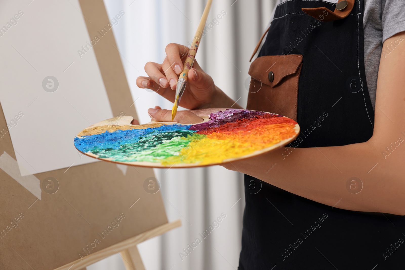 Photo of Woman mixing paints on palette with brush near easel in studio, closeup