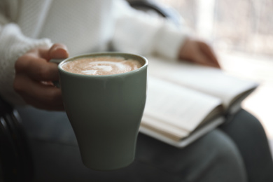 Woman with cup of coffee reading book indoors, closeup