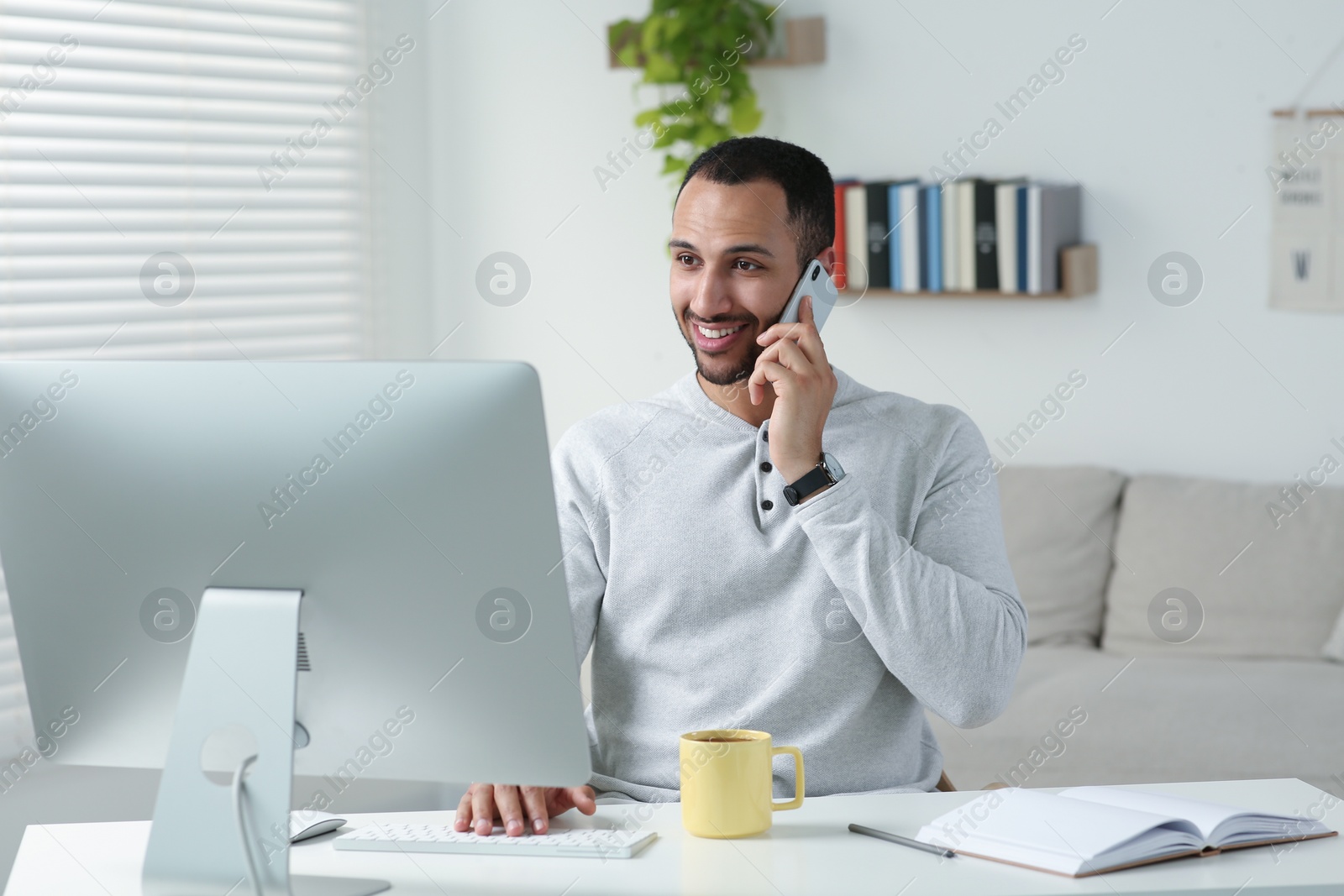 Photo of Young man talking on smartphone while working with computer at desk in room. Home office