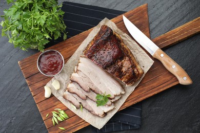 Photo of Pieces of baked pork belly served with sauce and parsley on black textured table, top view