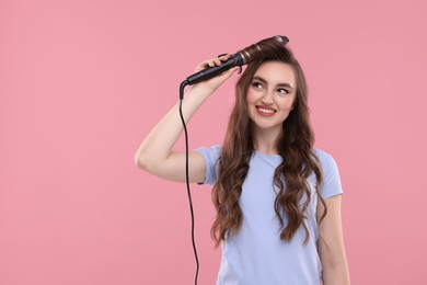 Beautiful young woman using curling hair iron on pink background, space for text