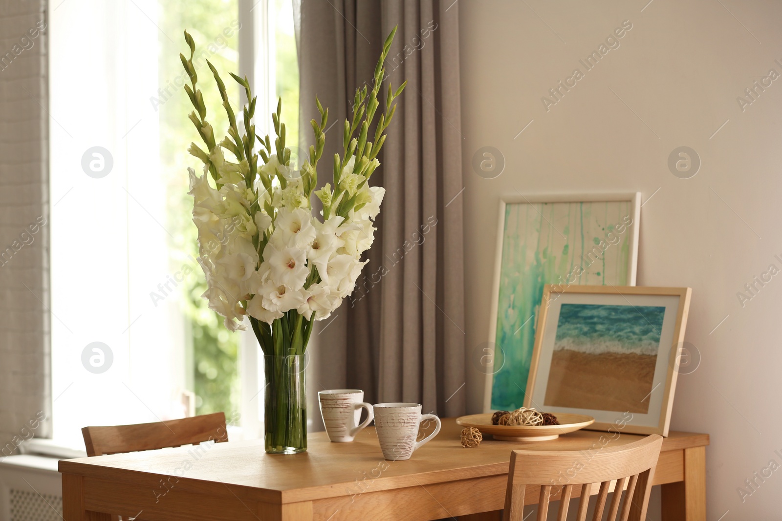 Photo of Vase with beautiful white gladiolus flowers, pictures and cups on wooden table in room, space for text