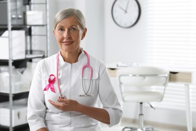 Photo of Doctor with stethoscope and pink ribbon indoors, space for text. Breast cancer awareness
