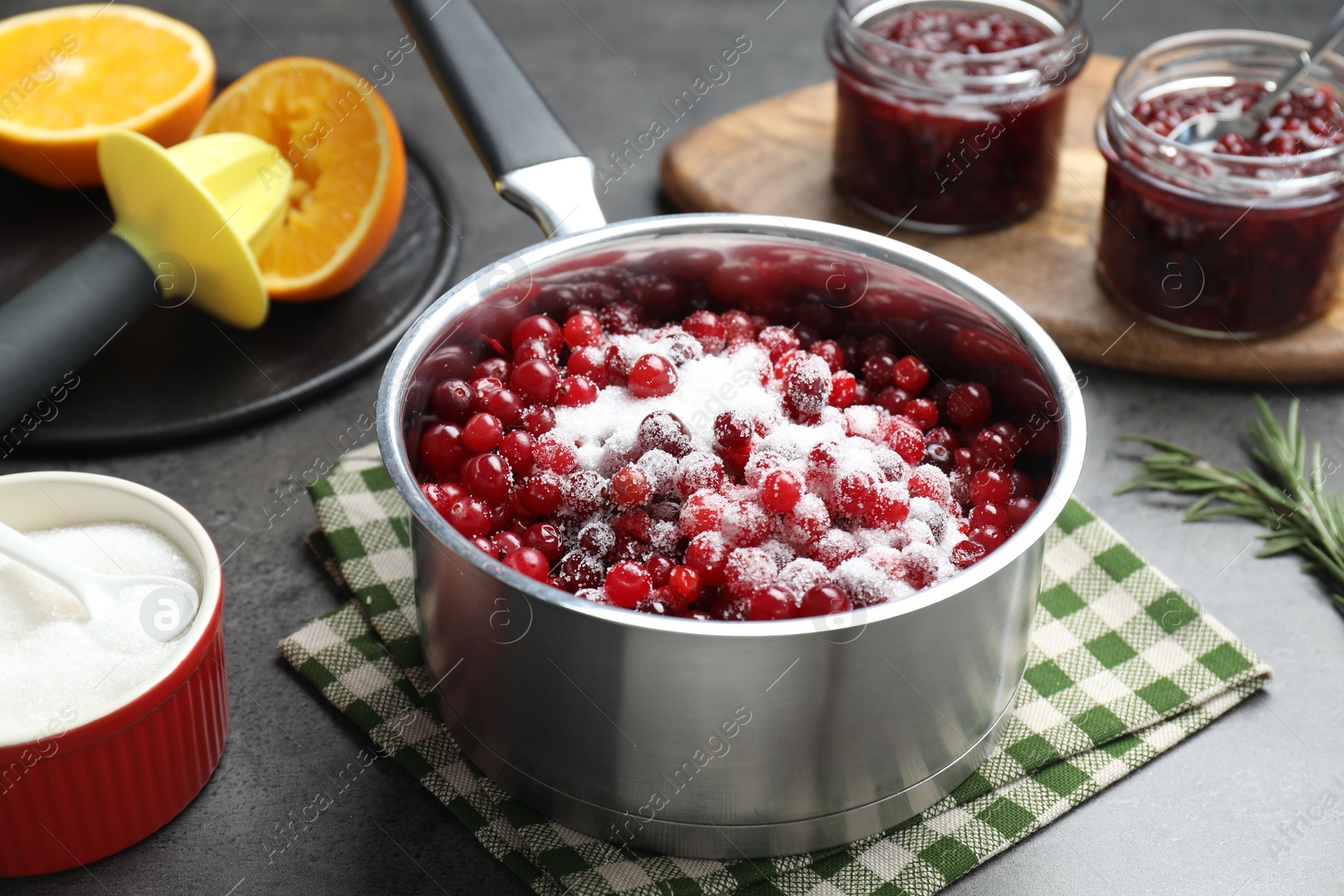 Photo of Making cranberry sauce. Fresh cranberries with sugar in saucepan and ingredients on gray table, closeup