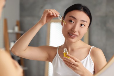 Beautiful young woman applying cosmetic serum onto her face near mirror in bathroom