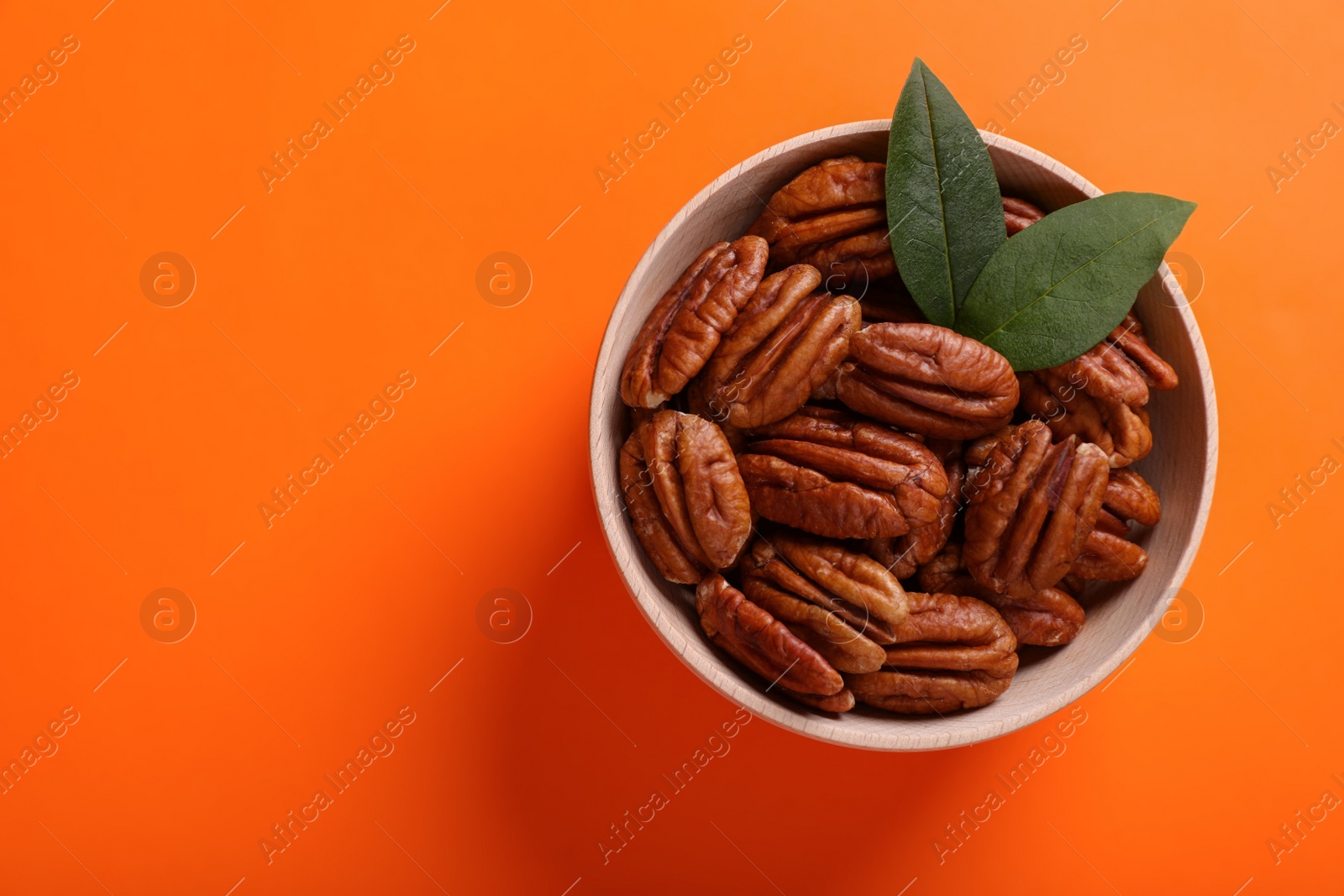 Photo of Bowl with tasty pecan nuts and green leaves on orange background, top view. Space for text