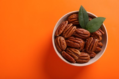 Photo of Bowl with tasty pecan nuts and green leaves on orange background, top view. Space for text