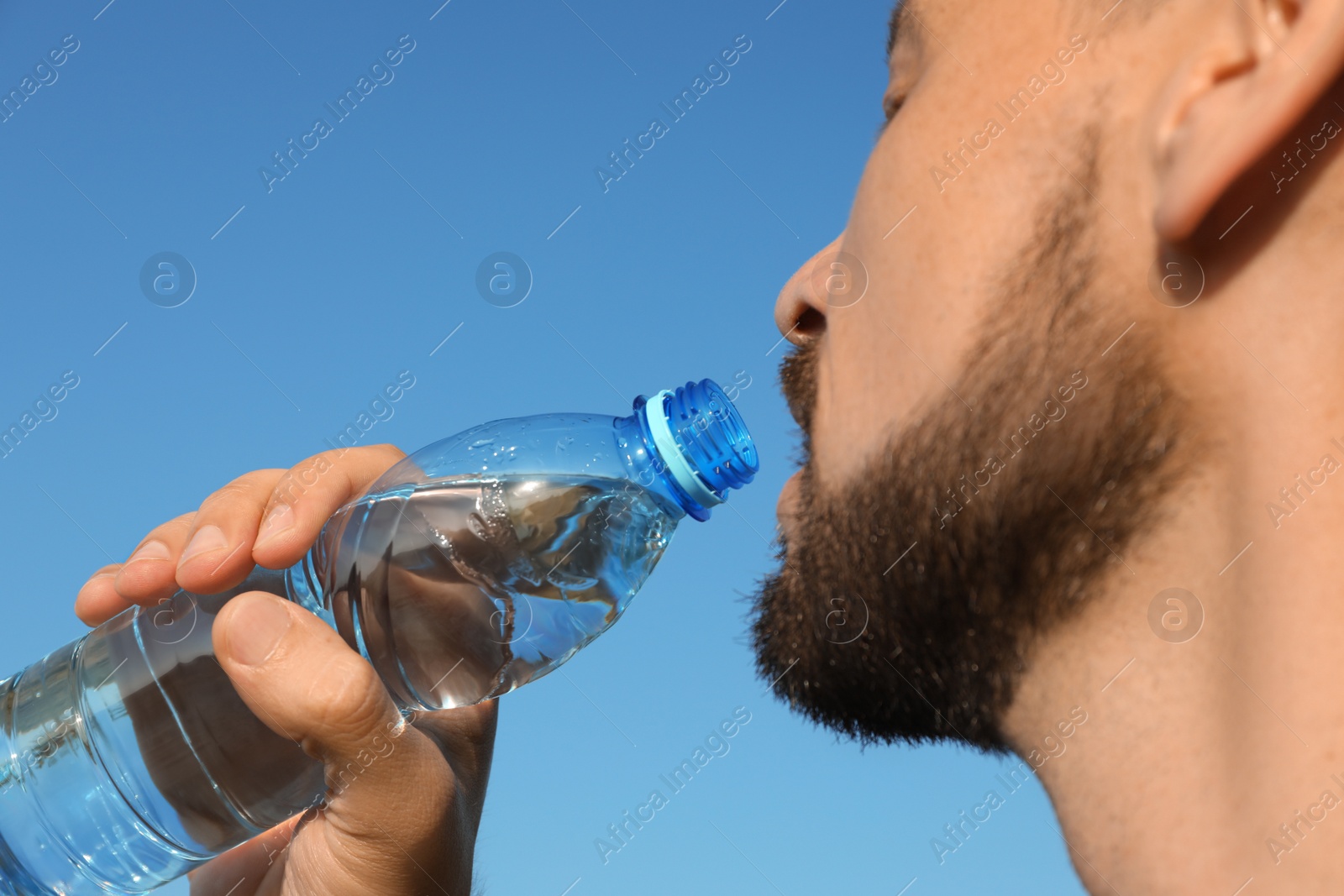 Photo of Man drinking water against blue sky on hot summer day, closeup. Refreshing drink