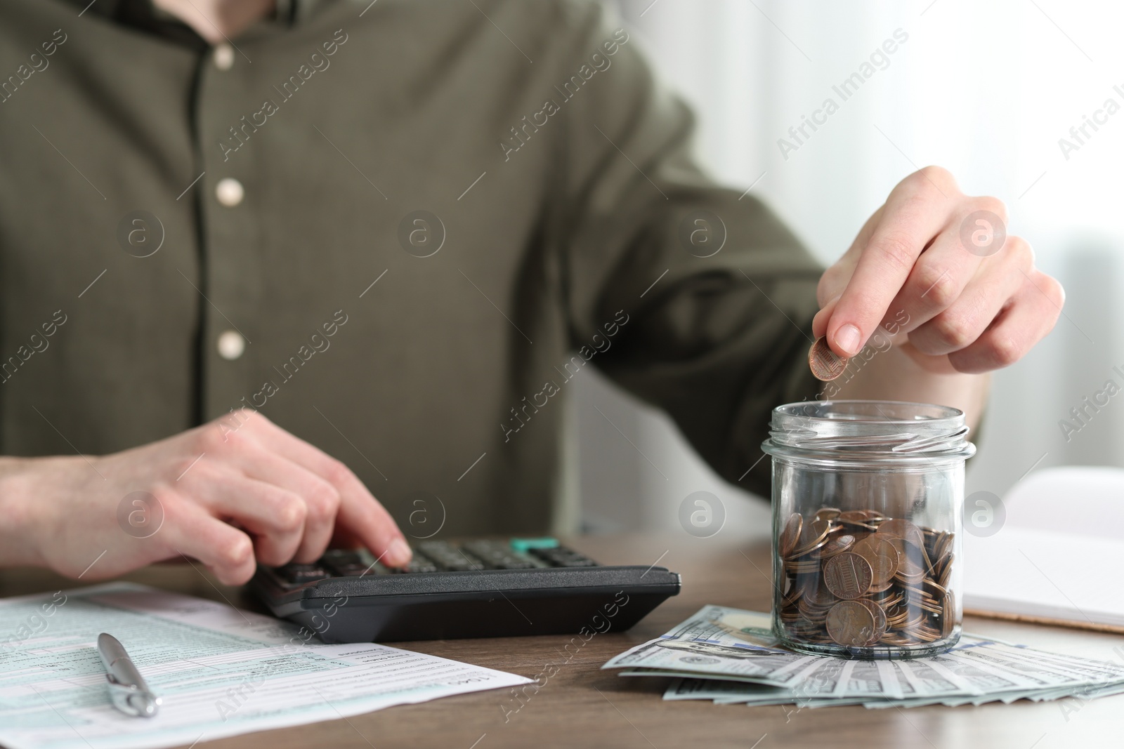 Photo of Financial savings. Man putting coin into glass jar while using calculator at wooden table, closeup