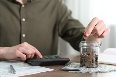 Financial savings. Man putting coin into glass jar while using calculator at wooden table, closeup