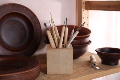 Photo of Set of different crafting tools and clay dishes on wooden rack in workshop