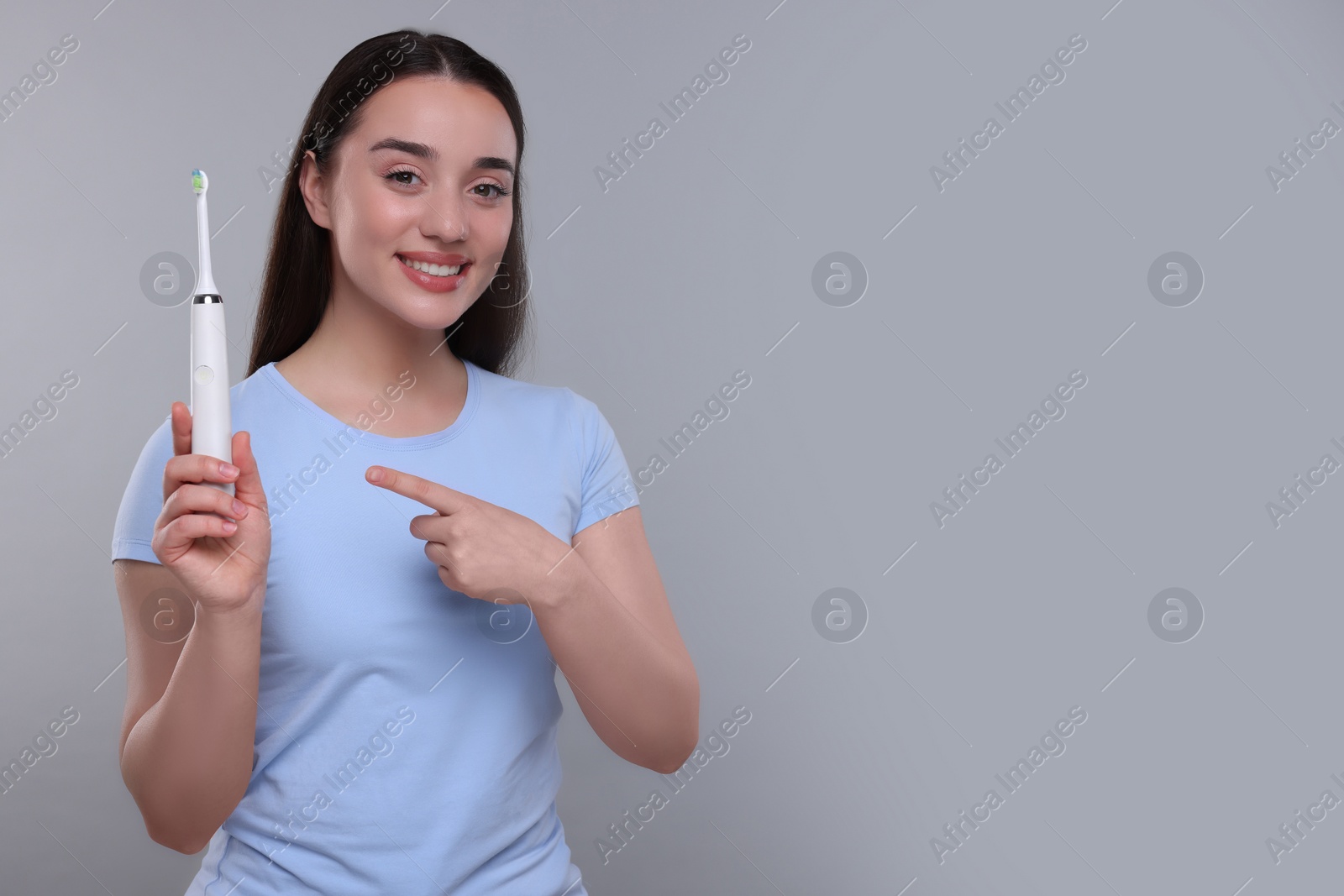 Photo of Happy young woman holding electric toothbrush on light grey background, space for text