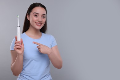 Happy young woman holding electric toothbrush on light grey background, space for text