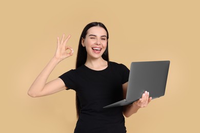 Photo of Happy woman with laptop winking and showing okay gesture on beige background